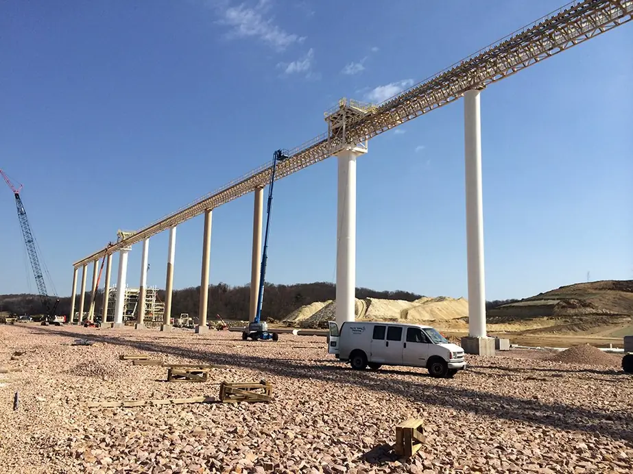 Sand plant conveyor gantry on tall pale-painted piers with a blue boom lift and a white van in the gravel yard