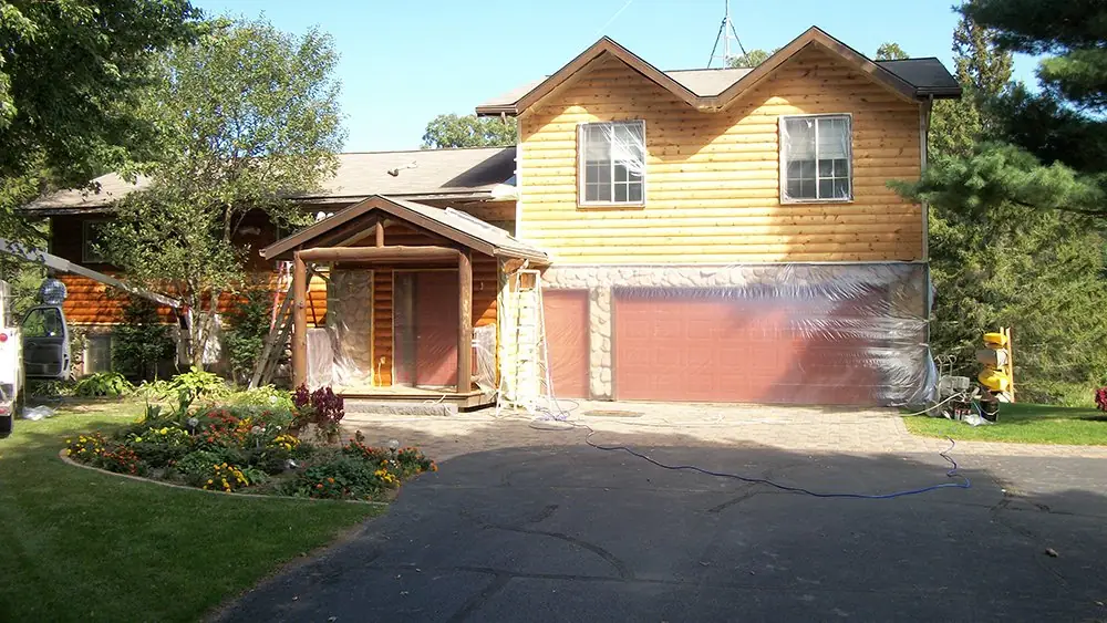 Log-style home with its garage wrapped in plastic sheeting while the walls are prepared for coating